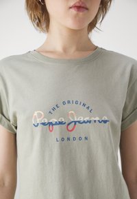Ljusgrön bomullströja med rund hals, korta ärmar och en flerfärgad tryckt logotyp: "THE ORIGINAL Pepe Jeans LONDON" centrerad.