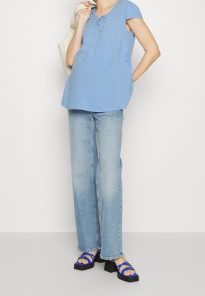 Persoon draagt een lichtblauwe blouse met korte mouwen, lichtblauwe spijkerbroek en blauwe sandalen met hak, draagt een beige tas over één schouder.
