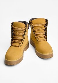 Gelbe Wildlederschuhe mit braunen Akzenten, runden Schnürsenkeln und Metallösen. Sie verfügen über eine dicke Gummisohle mit Profil und ein mittelhohes Design.