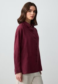 Camicia di lino borgogna con chiusura a bottoni, colletto e maniche lunghe con polsini a bottone, abbinata a pantaloni chiari.