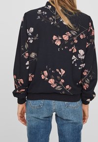 Svart blus med blommönster, som har rosa och vita blommor, långa ärmar, ribbade muddar och avslappnad passform. Tyget verkar lätt och texturerat.
