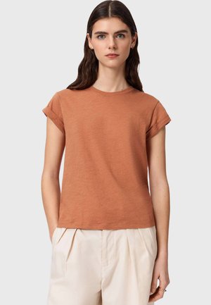 Młoda kobieta o długich ciemnych włosach nosi krótki, rdzawy rękaw i wysokie beżowe spodnie na jednolitym tle.