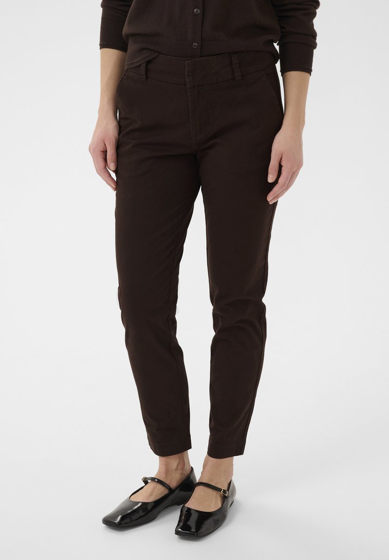Personne portant un pantalon slim foncé, un haut assorti foncé et des chaussures Mary Jane en cuir verni noir sur un fond clair uni.