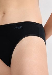 Close-up van een persoon in zwarte Sloggi onderbroek, met de nadruk op de taille en bovenbeen tegen een neutrale achtergrond.