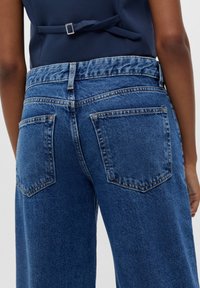 Jean en denim bleu taille haute avec une jambe évasée, comportant deux poches arrière et une finition de tissu texturé. Couture visible le long des coutures.