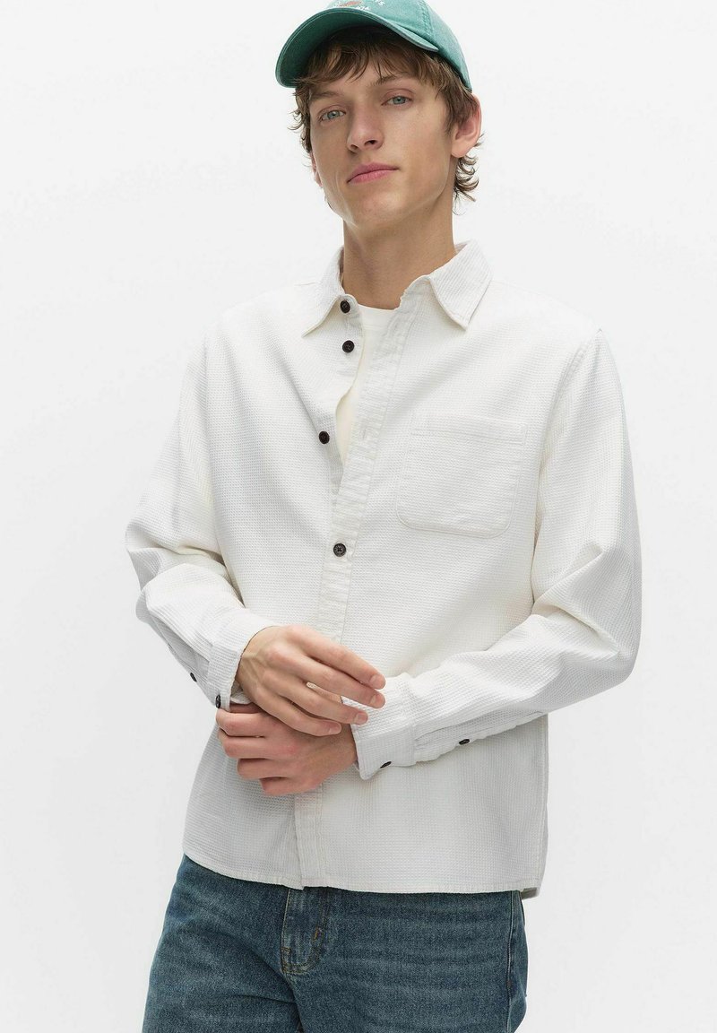 Jonge man met een lichtgroene pet, wit gestructureerd overhemd met knopen en voorzak, en blauwe spijkerbroek tegen een effen witte achtergrond.