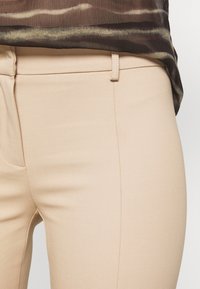 Beige fitted trousers with a smooth texture, featuring a slender waistband and subtle side pockets, partially covered by a patterned top.