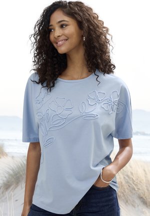 Lächelnde Frau mit lockigem Haar, die ein hellblaues T-Shirt mit Blumenstickerei trägt und an einem Sandstrand mit Gras und Meer im Hintergrund steht.