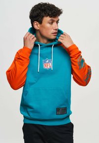 Hoodie i turkos och orange. Har NFL-logotyp på bröstet, kenguruficka och grafik på ärmen. Mjuk tygkänsla.