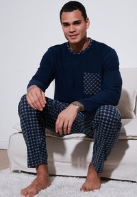 Mornarska pidžama sastavljena od majice s dugim rukavima koja ima karirani džep, u kombinaciji s kariranim hlačama tamnih nijansi. Mekani materijal i opušten kroj.