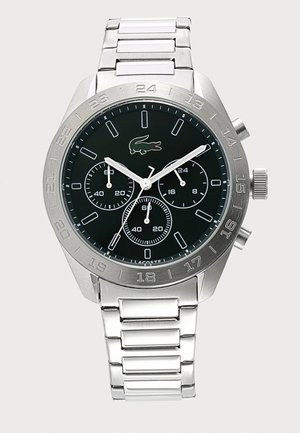 Montre-bracelet en métal argenté avec cadran noir, trois petits cadrans, index des heures blancs et logo crocodile à la position de 12 heures.