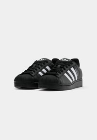Black leather sneakers with a textured rubber toe cap, featuring three white stripes and logo on the tongue. Classic low-top design.