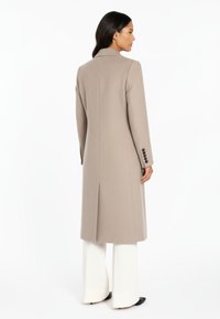 Beige ullkappa med skräddarsydd passform, långa ärmar och en slits baktill. Svarta knappluckor på ärmarna, kombinerade med vita vida byxor.