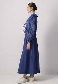 Blusa blu a fantasia con maniche a sbuffo e nodo davanti, abbinata a una lunga gonna blu. Sneakers bianche completano l'outfit.