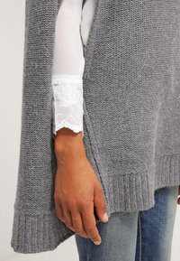 Grå stickad poncho med ribbad nederkant, med en split-design som överlappar en vit spetsskjorta vid ärmen med blommönster.