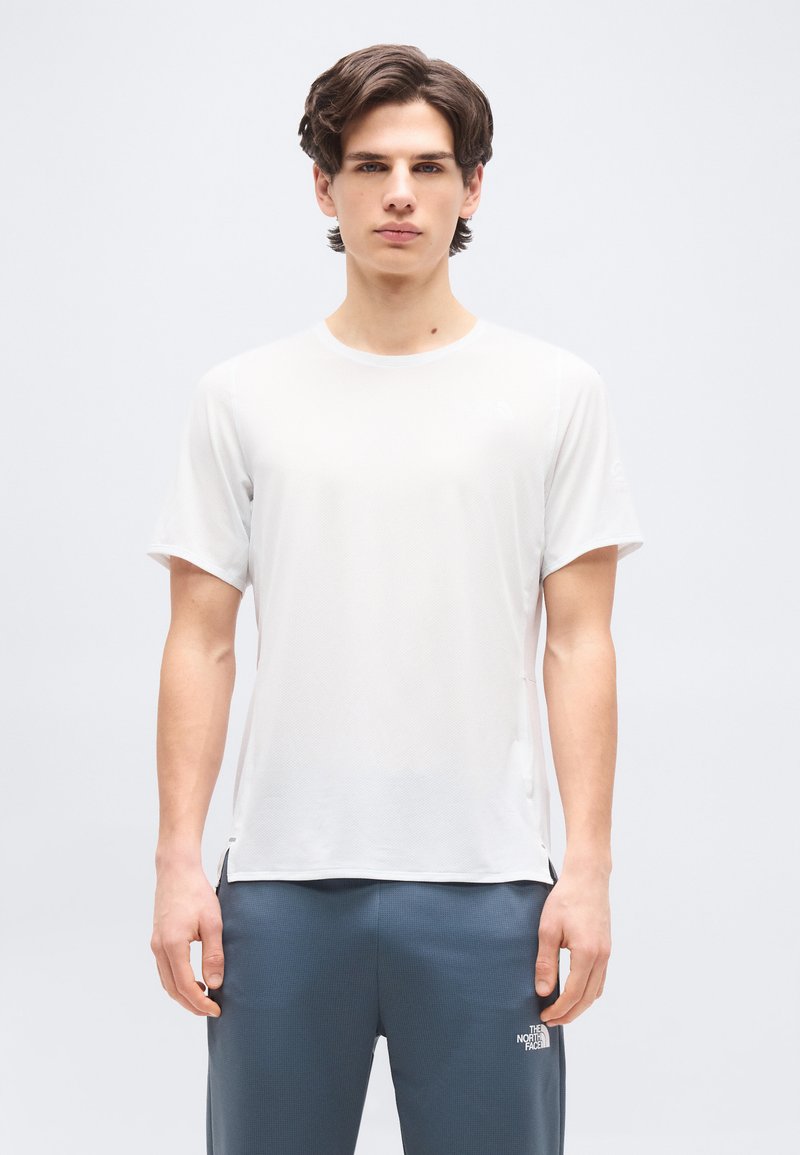 Νεαρός άνδρας με κοντά καστανά μαλλιά που φοράει ένα απλό λευκό κοντομάνικο πουκάμισο και γκρι παντελόνι με λογότυπο στο αριστερό μηρό.