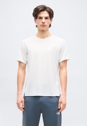 Νεαρός άνδρας με κοντά καστανά μαλλιά που φοράει ένα απλό λευκό κοντομάνικο πουκάμισο και γκρι παντελόνι με λογότυπο στο αριστερό μηρό.