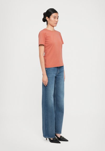 Kvinna som står på sidan i en rostfärgad kortärmad skjorta, vidbeniga blåjeans och svarta spetsskor mot vit bakgrund.
