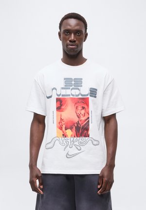 Męski model noszący białą koszulkę Nike z napisem „Be Unique” i czerwonym nadrukiem osoby wskazującej, w połączeniu z ciemnymi szortami.