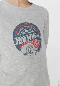 Šedá dlouhá tričko s dlouhým rukávem, které má opotřebované logo Hot Wheels s grafikou kola a plamene v červené a modré barvě. Bavlněný materiál, design s kulatým výstřihem.