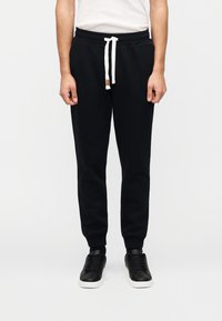 Μαύρο βαμβακερό παντελόνι φόρμας με στενό σχεδιασμό, ελαστική μέση, άσπρο κορδόνι και ριμπ τελείωμα, σε συνδυασμό με μαύρα αθλητικά παπούτσια.