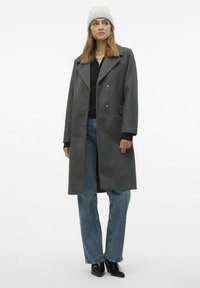 Cappotto lungo grigio in misto lana con colletto a punte, chiusura doppiopetto, tasche laterali, indossato sopra un top nero e jeans blu, abbinato a un berretto lavorato a maglia.