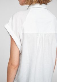 Camicia bianca in lino a maniche corte con colletto, dotata di uno spacco sul retro e polsini arrotolati, che mette in mostra una consistenza morbida e un aspetto leggero.