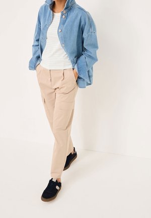 Person iført beige cargobukser, hvid stribet skjorte, lyseblå cowboyjakke og sorte sneakers, stående med hænderne i lommerne.