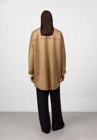 Persoon met lang bruin haar, gekleed in een beige oversized jas en zwarte wijde broek, staand tegenover een simpele witte muur.
