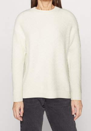 Femme portant un pull en maille texturé blanc à manches longues associé à un jean gris foncé, debout devant un fond clair uni.