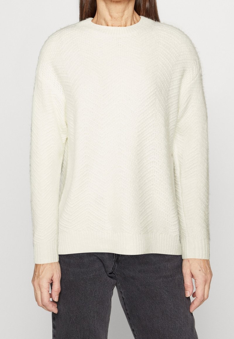 Femme portant un pull en maille texturé blanc à manches longues associé à un jean gris foncé, debout devant un fond clair uni.