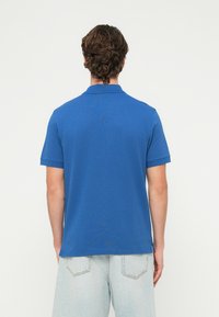 Άνδρας με καστανά κατσαρά μαλλιά, φορώντας μπλε πόλο μπλουζάκι με κοντά μανίκια και ανοιχτόχρωμο τζιν σορτς, φαίνεται από πίσω σε απλό φόντο.