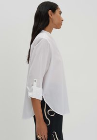 White long-sleeve shirt with a high collar, roll-up cuffs featuring snap detail, and a loose fit, paired with patterned black trousers.