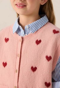 Enfant portant un gilet en tricot rose avec des motifs de cœurs rouges par-dessus une chemise à col rayée bleue et blanche.