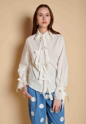 Young woman wearing a white blouse with bows down the front and at cuffs, paired with blue pants featuring large white polka dots.