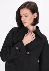 Schwarze, strukturierte Jacke mit Kragen, Knopfverschluss und gerollten Ärmeln, die mit silberfarbenen Knöpfen verziert sind. Glattes Material ohne auffällige Muster.