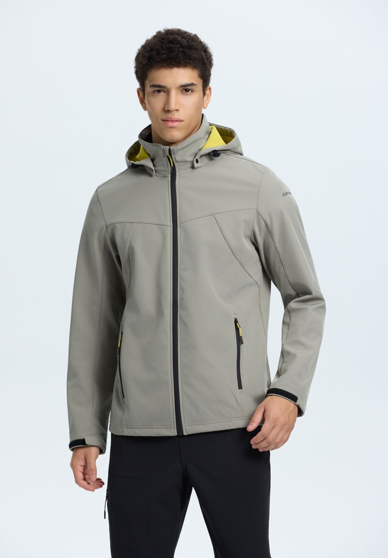 Veste softshell gris clair avec une capuche, des fermetures éclair noires et une doublure jaune. Présente un design élégant et plusieurs poches zippées.
