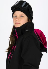 Giacca da sci nera con accenti rosa, colletto alto e zip visibili. Indossata con un berretto nero e occhiali da sci appoggiati sulla fronte.