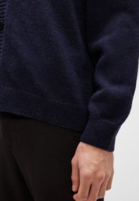 Kék kötött kardigán texturált felülettel, bordázott aljjal és mandzsettával, minimalista logóval az alsó szélén.