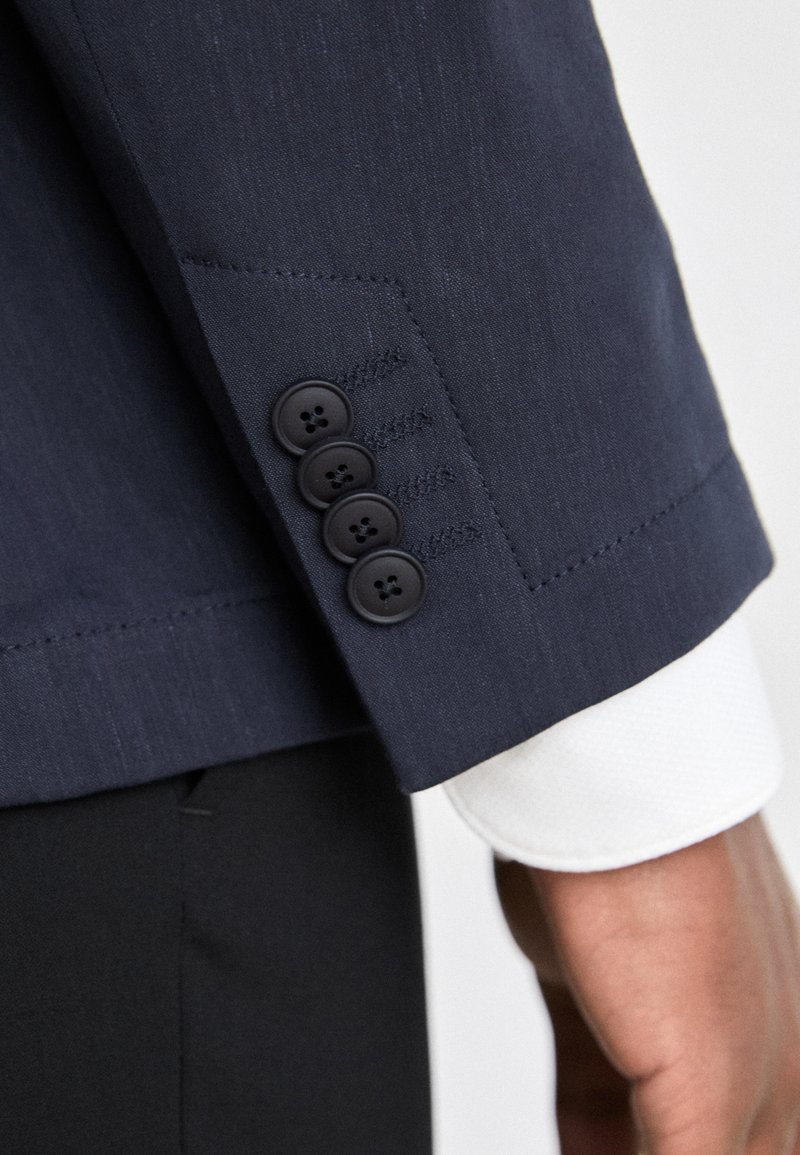 Manteau de costume marine avec des poignets à quatre boutons noirs, tissu texturé subtil, et le poignet d'une chemise blanche qui dépasse en dessous, mettant en valeur le design sur mesure.