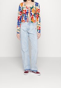 Bunt gestreifter Langarmtop mit floralem Muster und vorderem Knoten, kombiniert mit hellblauen geraden Jeans und kastanienbraunen Sneakers.