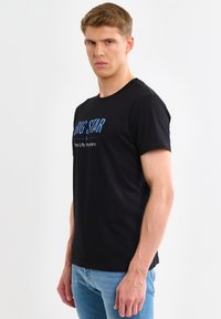 Giovane uomo con T-shirt nera "BIG STAR" e jeans azzurri, in piedi di lato contro uno sfondo bianco semplice.