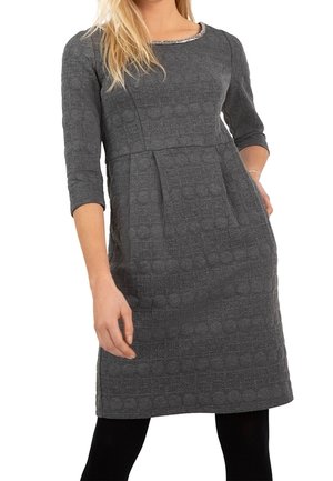 Femme portant une robe grise texturée arrivant au genou avec des manches trois-quarts, debout avec une main sur la hanche, leggings noirs visibles.