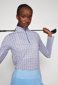 Langermet golftrøye med kvart glidelås, med et geometrisk mønster i rosa, lilla og blå på en lys blå bakgrunn, med en jevn tekstur.