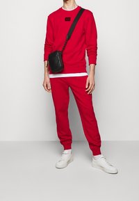 Personne portant un sweat-shirt rouge et un pantalon de survêtement assorti, des baskets blanches et un sac bandoulière noir, se tenant devant un fond clair uni.