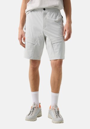 Man die lichtgrijze cargoshorts, witte sportsokken en grijze sneakers met oranje accenten draagt, staand tegen een effen achtergrond.