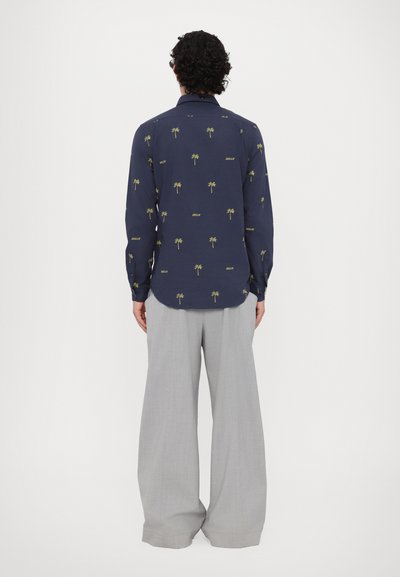 Camicia a maniche lunghe blu navy con stampa gialla di una palma e "DREAM", abbinata a pantaloni larghi grigi chiari. Vista posteriore, tessuto liscio.
