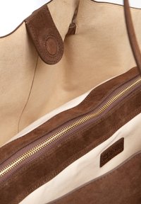 Brązowa zamszowa torba typu tote z beżową podszewką wewnętrzną, wyposażona w kieszeń na suwak ze złotymi okuciami oraz małą naszywką z logo.