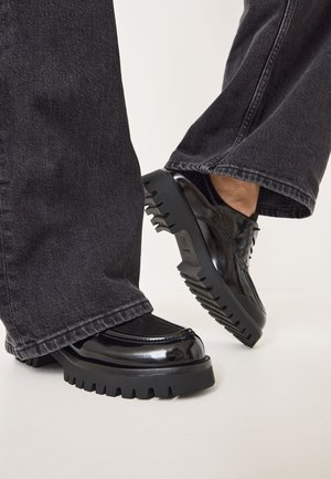 Scarpe in pelle verniciata nera con suola spessa, caratterizzate da cuciture decorative e punta arrotondata. Indossate con pantaloni in denim scuri.