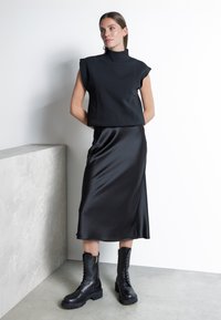 Svart ärmlös topp med hög hals, kombinerad med en svart satin midi-kjol. Modellen bär svarta snörboots med en grov sula.